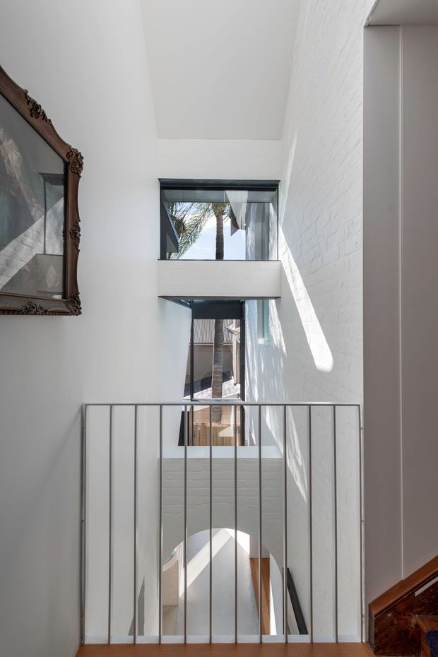 Bright, narrow double-height interior stairwell or landing with white-painted brick walls, a simple grey metal balustrade, timber flooring, and tall black-framed windows bringing in strong daylight. A partial ornate framed artwork and timber stair details add traditional contrast to the otherwise clean, contemporary space. The view looks across an open void toward multiple levels and exterior greenery beyond the glazing.