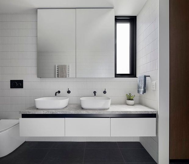 Contemporary double-vanity bathroom with a wall-mounted white vanity, two basins and black fixtures. Fine vertically gridded wall tiles create a clean backdrop, while a dark tiled floor, grey stone-look benchtop, mirrored wall cabinets and a narrow black-framed window add contrast.