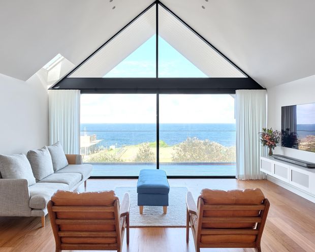 Bright coastal living room with a dramatic triangular gable window framing an ocean view. The room includes a pale upholstered sofa, two mid-century wooden lounge chairs, a blue ottoman, light timber flooring, white curtains, and a white built-in media cabinet. The high pitched glazed wall and abundant daylight create an airy, refined atmosphere.