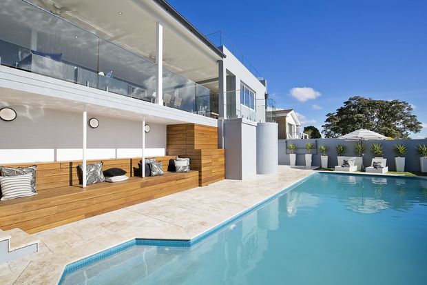 Bright exterior view of a contemporary multi-level residence with a large swimming pool, expansive upper-level glazing, glass balustrades, white rendered walls, and warm timber-clad built-in poolside seating. The pool terrace includes outdoor lounge furniture and raised planters, with a clear blue sky and mature tree beyond.