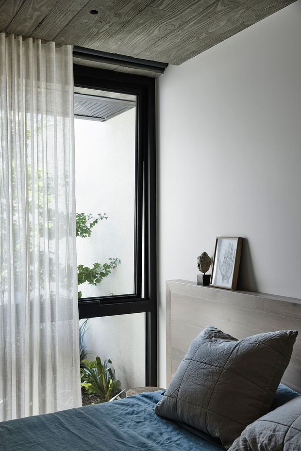 A calm, contemporary bedroom corner with a bed in the foreground, a pale timber-look headboard, charcoal bedding and a large black-framed window beside sheer curtains. The room has a textured wood ceiling, white walls, and a small framed artwork and decorative object on the headboard shelf. A planted courtyard is visible through the window.