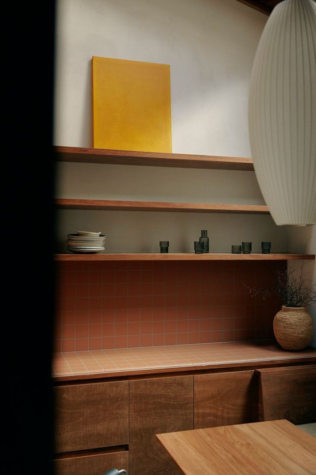 A warm, moody interior detail showing a built-in timber sideboard or dining-area storage unit with open shelving. The shelves display ceramics and small objects, while the central recessed surface is lined with terracotta-red square tiles and a matching tiled counter. A large ribbed pendant light hangs at the right, with a woven vase and branches below. A timber dining table is visible in the foreground.
