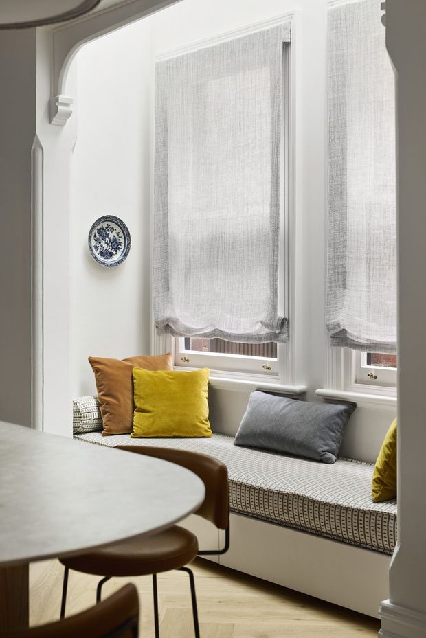 A refined dining nook with a built-in window seat beneath two tall windows. The seat has a patterned upholstered cushion and mustard, ochre, and grey throw pillows. A round dining table and curved brown chairs occupy the foreground. The space features white painted trim and joinery, light timber flooring, soft grey walls, sheer textured roller blinds, and a decorative blue-and-white wall plate. The overall look is bright, warm, and traditionally detailed.