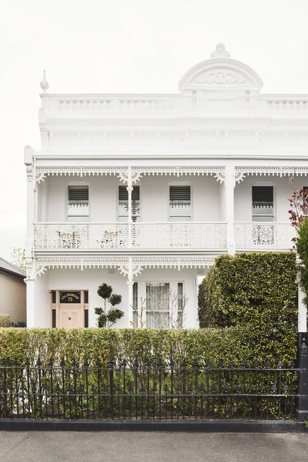 Front elevation of a narrow two-storey heritage terrace or townhouse with a white rendered facade, ornate cast-iron lacework verandah and balcony railings, decorative brackets, classical detailing, tall shuttered windows, and a formal hedge behind a black metal fence. The image is an exterior architectural view with a restrained white and green palette.