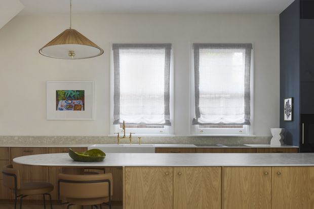 Warm, refined kitchen interior with a long central island, natural timber cabinetry, pale stone benchtops, brass-toned tapware, and two softly screened windows. A woven pendant light, framed artwork, and timber-framed stools add understated character. Dark full-height cabinetry or wall joinery appears at the right edge.