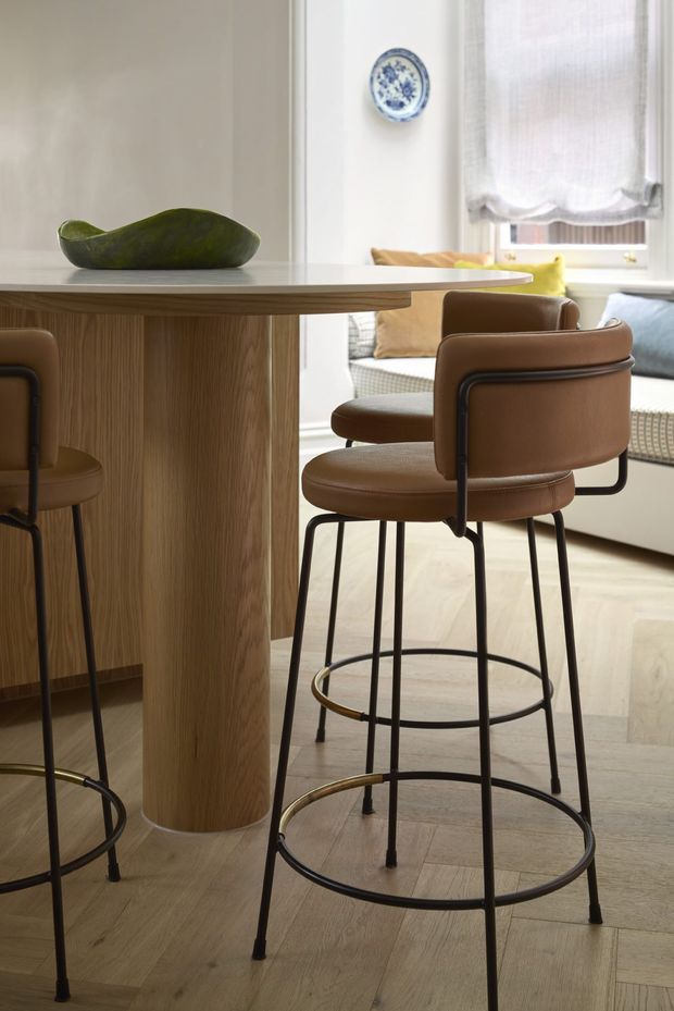 A refined open-plan dining and living area centered on a round light-wood kitchen or dining island with a thick pale benchtop. Brown upholstered bar stools with slim black metal frames surround the island. Light timber flooring, white walls, a large window with a soft Roman blind, and a built-in window seat with cushions create a bright, warm interior. A decorative blue-and-white plate and sculptural green bowl provide restrained accents.