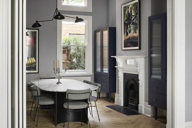 A refined dining room with a round marble dining table, upholstered dining chairs, dark built-in display cabinetry, a decorative traditional fireplace, tall sash windows, and herringbone timber flooring. The room combines classic architectural details with contemporary dark joinery and sculptural black pendant lighting.