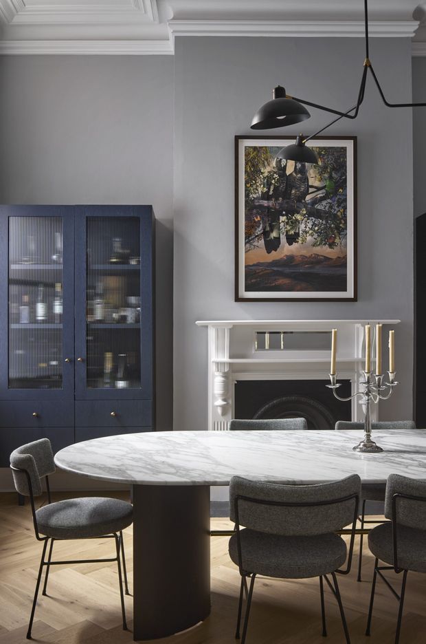 Elegant dining room with a large oval white marble dining table, upholstered charcoal dining chairs, dark blue built-in glass-front cabinetry, a decorative white fireplace mantel, framed artwork, and sculptural black pendant lighting. The room has grey walls, ornate white ceiling cornices and mouldings, and a light timber parquet floor, creating a refined contemporary-traditional interior.