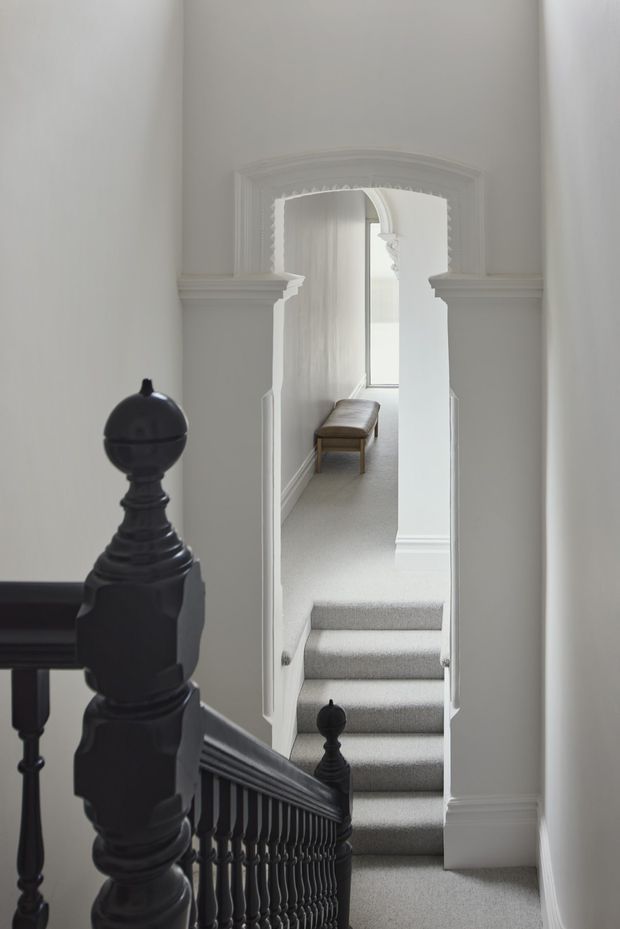 A softly lit interior view of a traditional stairway and hallway. Dark stained timber balustrades and turned newel posts contrast with white walls, ornate moulded architraves, decorative archway detailing, and pale carpeted stairs. The stair rises toward a narrow landing with a small upholstered bench and a tall window beyond.