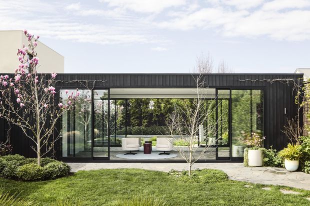 Exterior view of a modern, single-storey pavilion-like building with a dark vertically ribbed facade and extensive floor-to-ceiling glazing. The transparent living area opens visually onto a landscaped garden, with minimalist outdoor paving and planted borders. A white geometric volume is visible behind the pavilion, and the flat roofline gives the composition a clean contemporary character.