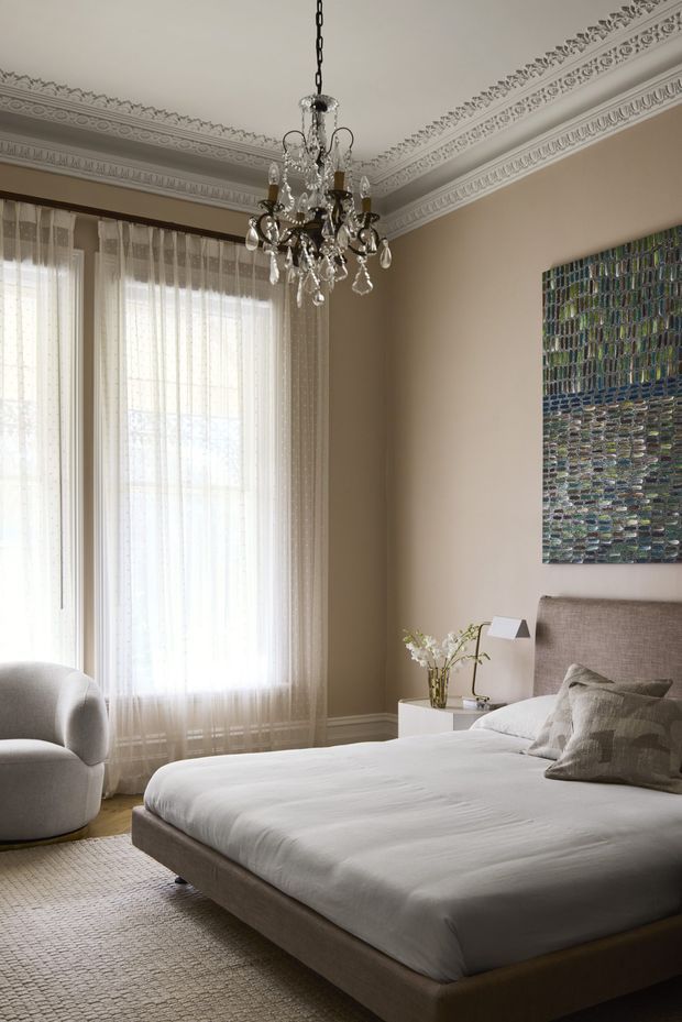 Elegant bedroom with a low upholstered bed, neutral bedding, textured grey-beige headboard, curved armchair, sheer floor-to-ceiling curtains, ornate decorative ceiling cornices, and a crystal chandelier. A colourful abstract artwork adds contrast to the otherwise refined, warm neutral scheme.
