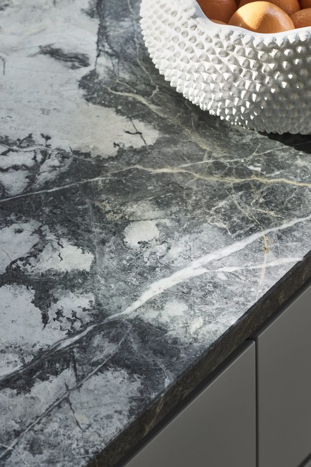 Close-up of a dark grey, heavily veined stone benchtop with a thick edge profile, paired with smooth dark grey fixed cabinetry. A white textured bowl containing oranges sits on the surface. The image focuses on the countertop material and built-in joinery rather than a complete room.