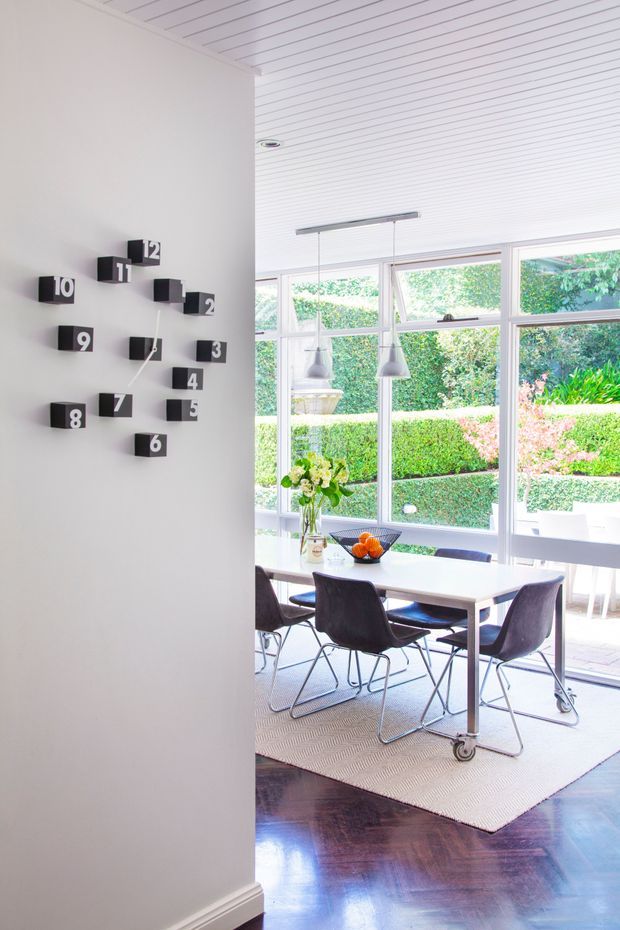 Bright, contemporary dining area viewed from an adjacent hallway or entry space. A white rectangular dining table is surrounded by dark sculptural chairs, with pendant lights above. Floor-to-ceiling glazing opens to a manicured garden, while a sculptural black wall clock decorates the white wall. The room features a white slatted ceiling, glossy dark timber flooring, and a large neutral rug.