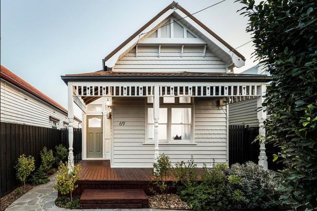 Front exterior view of a compact heritage-style weatherboard cottage with a steep gabled roof, decorative white fretwork veranda, timber steps and a landscaped entry garden. The facade features horizontal cladding, white trim, a glazed front door and sash-style windows.
