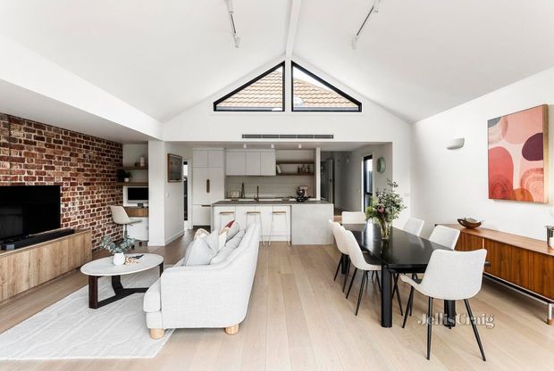 Bright open-plan living, dining and kitchen interior with a vaulted white ceiling and triangular clerestory windows. The room features a red exposed-brick feature wall, light timber flooring, white cabinetry and furnishings, a black dining table, a timber media console, and colourful abstract wall art.