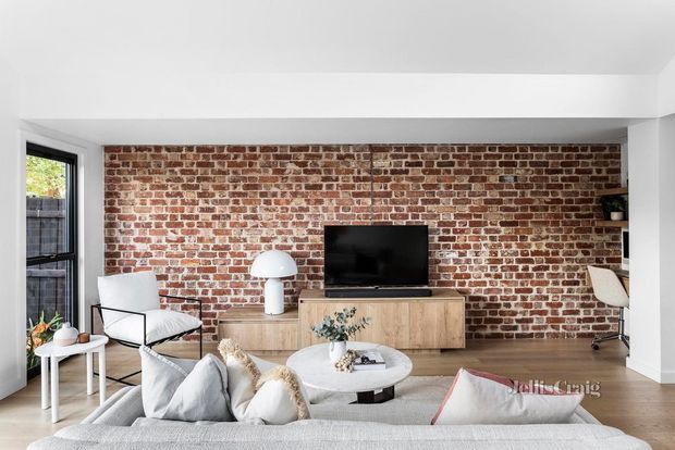 A contemporary living room with a full-width exposed red-brick feature wall, white ceiling and trim, timber flooring, and a low timber media console. The room includes a large sectional sofa, round coffee table, modern lounge chair, sculptural table lamp, television, and a small home office nook visible at the right. Natural light enters through the tall window at left.