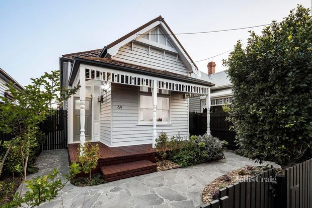 Front exterior view of a compact heritage-style weatherboard cottage with a steep gabled roof, decorative veranda, timber posts and fretwork, small front deck, and irregular grey stone paving. The facade is painted pale grey with white trim, surrounded by clipped shrubs and black fencing.
