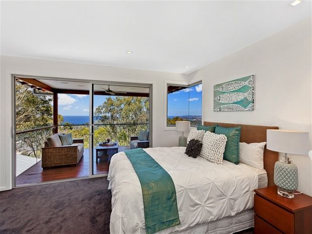 Bright, coastal-inspired bedroom with a large bed dressed in white bedding and a turquoise runner, layered teal and textured accent pillows, timber bedside tables, and matching lamps. Sliding glass doors open to a balcony with leafy views, while framed aquatic artwork and ocean-blue tones reinforce the relaxed coastal character. The room has a white ceiling and walls, dark plush carpet, and abundant natural light.