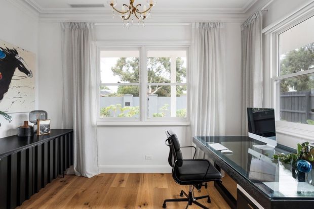 Bright, refined home office with white walls, ornate ceiling cornices, full-length sheer curtains, warm timber flooring, and large windows overlooking a garden. A glass-topped dark desk with computer and ergonomic chair occupies the right side, while a black ribbed sideboard and artwork line the left wall. A small chandelier adds a traditional decorative accent.