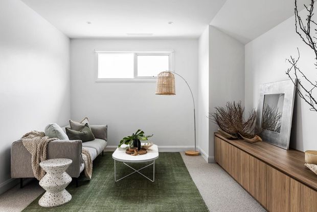 A bright, compact living room with white walls and ceiling, a grey upholstered sofa layered with neutral throws and cushions, a green area rug, and a round white coffee table. A slim arched floor lamp, potted plant, sculptural side table, and wood-grain built-in media console with mirror and dried branches add warmth and texture. The room has a calm, contemporary minimalist aesthetic with a small high window and recessed ceiling lights.