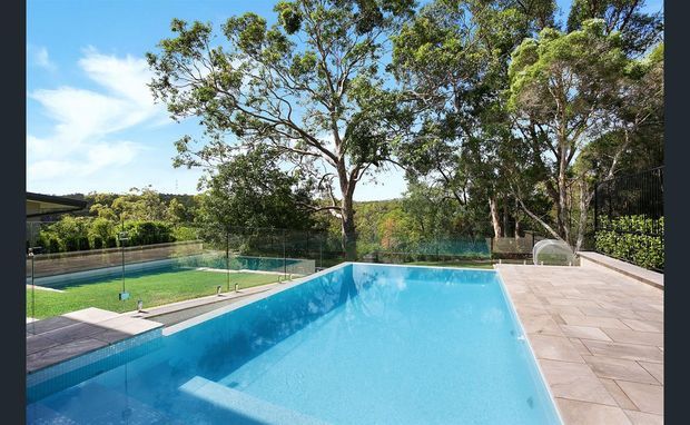 Outdoor swimming pool with bright blue water, surrounded by pale tiled paving and transparent safety fencing. Mature trees and landscaped greenery form the backdrop, with a second lawn and pool area visible beyond.