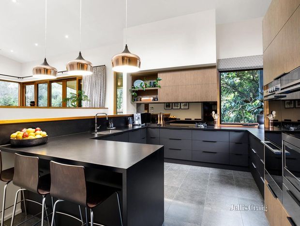 A contemporary kitchen with a large black island and integrated breakfast bar, dark cabinetry, warm timber overhead joinery, and a black benchtop. The kitchen features pendant lighting, integrated appliances, grey tiled flooring, and extensive windows overlooking greenery.