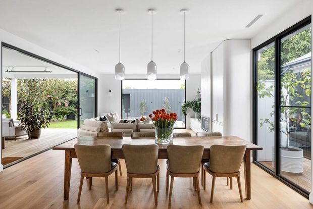 Bright open-plan dining and living space with a timber dining table, six upholstered dining chairs, a neutral sectional sofa, and three pendant lights. Full-height black-framed glazing opens the room to a landscaped courtyard, while white built-in cabinetry and light timber flooring create a clean contemporary interior.
