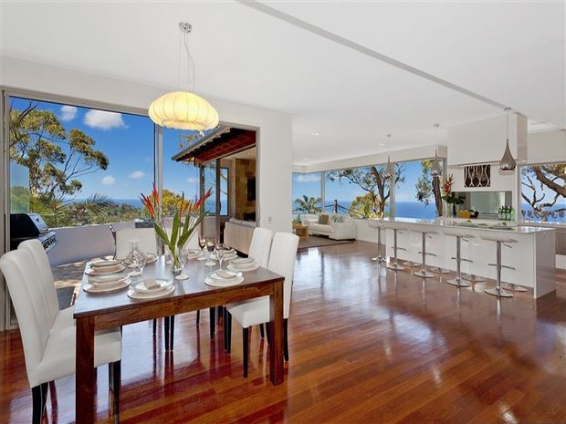 Bright open-plan interior combining a formal dining area and contemporary kitchen, with polished timber flooring, white cabinetry, expansive glazing and ocean views. The dining table is set for a meal, while a white kitchen island with bar seating extends into the living area. A balcony or terrace and landscaped coastal surroundings are visible beyond the glazing.