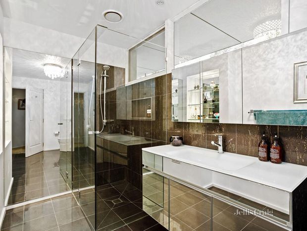 A spacious contemporary bathroom with a long double vanity, white integrated basins, mirrored cabinetry, a frameless glass walk-in shower, and dark brown stone-look wall and floor tiles. Bright white walls and ceiling, extensive mirrors, chrome fixtures, and glass shelving create a polished, reflective interior.