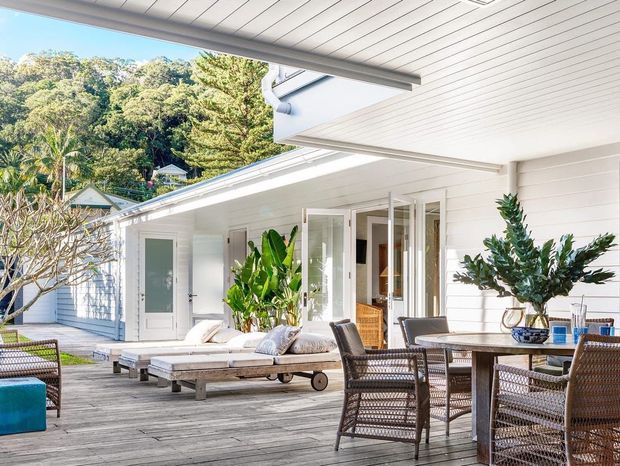 Bright, relaxed outdoor entertaining area beside a white weatherboard home, featuring a spacious timber deck, dining table and woven chairs, sun loungers, tropical plants, and wide-opening glazed doors. Lush wooded surroundings are visible beyond the property.