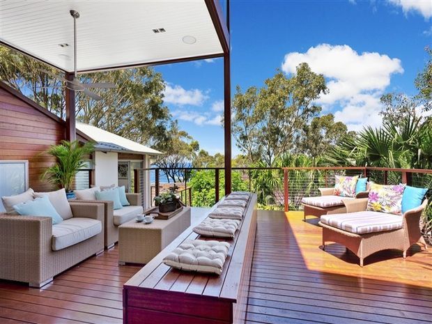A spacious elevated outdoor terrace or deck furnished as an outdoor living and lounge area. The space features timber decking, a white-lined covered ceiling, warm timber and red-toned railings, wicker sofas and armchairs with pale cushions, a long built-in timber bench with tufted cushions, potted palms, and views through the railing toward trees and blue sky.