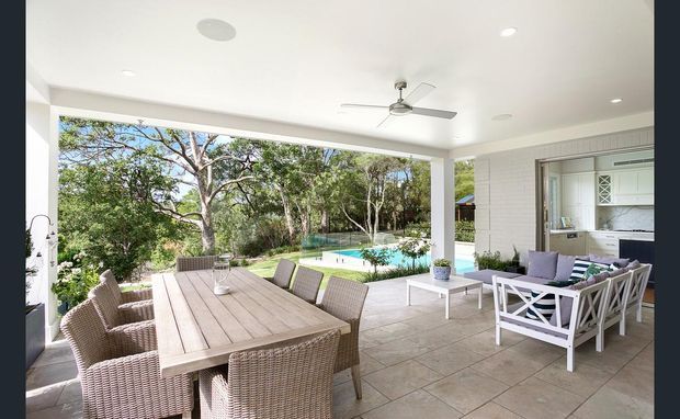 Covered outdoor entertaining terrace with a large timber dining table and wicker chairs, a white outdoor lounge setting, ceiling fan, tiled flooring, and a swimming pool surrounded by landscaped greenery beyond. The space is bright, open, and designed for indoor-outdoor living.