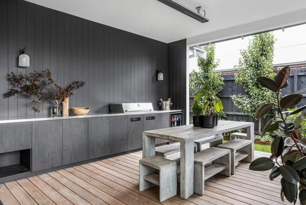 A contemporary covered outdoor entertaining area with a built-in dark kitchen and barbecue wall, set against vertical charcoal cladding. The space opens onto a timber deck and includes a substantial weathered timber dining table with bench seating, potted plants, and a fenced garden outlook. Minimal wall lights and a linear ceiling light complete the refined outdoor setting.