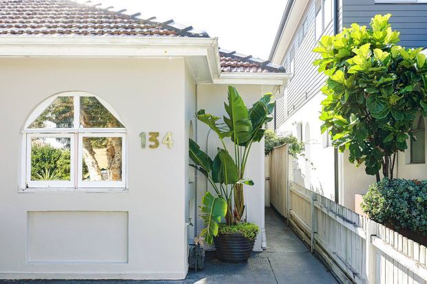 Bright exterior view of a narrow residential side passage between two closely spaced homes. The foreground house has a white rendered facade, arched-top window, recessed wall panel, pale tiled pitched roof, and the number 134 displayed beside the window. Tropical potted plants line the passage, with a timber fence and neighbouring weatherboard-clad building visible on the right.