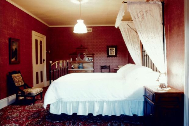 A traditional bedroom with a large white bed dressed in a ruffled bedspread, deep red patterned walls, a floral patterned carpet, lace canopy curtains, a cream beadboard ceiling, and dark wood furnishings. Decorative framed artwork, a small chair, dresser, and bedside table complete the richly furnished room.