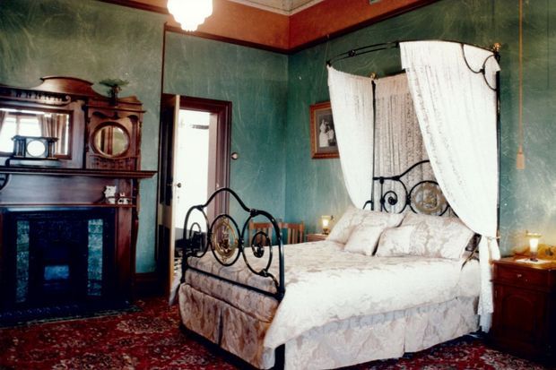 A richly decorated traditional bedroom with a black wrought-iron four-poster bed draped in white curtains, patterned bedding, dark wood bedside furniture, an ornate timber fireplace mantel, framed artwork, and a patterned area rug. The walls are deep teal-green with dark red-brown trim and decorative ceiling moulding.