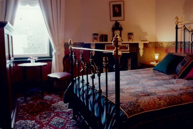 A richly furnished traditional bedroom with an ornate dark metal four-poster bed, patterned bedspread, jewel-toned cushions, heavy drapery, Persian-style rug, dark timber furniture, and a decorative fireplace mantel. Warm ambient lighting and antique detailing create an elegant, historic atmosphere.