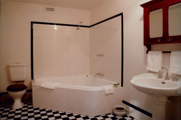 Traditional-style bathroom with a white tiled bath and shower enclosure, black trim, black-and-white checkerboard floor tiles, pedestal sink, toilet, and dark wood mirrored wall cabinet. The room has a high-contrast, period-inspired fit-out.