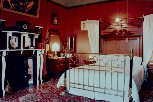 A richly decorated traditional bedroom with deep red walls, an ornate white fireplace and shelving, a brass-and-black metal four-poster bed, antique timber furniture, mirrors, framed artwork, and a patterned oriental-style carpet. White curtains and bedding contrast with the dark, dramatic colour scheme.