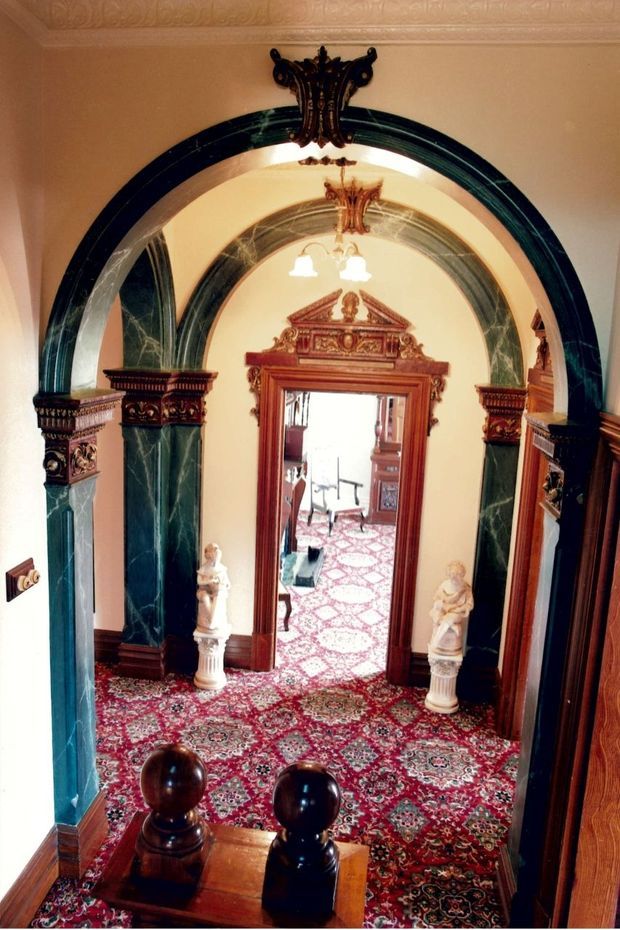 Ornate traditional interior hallway viewed from a staircase, featuring a richly patterned red carpet, dark timber balustrade and door surrounds, green marble-clad columns, elaborate carved archways, decorative plaster ceiling moulding, and classical statues flanking a doorway. The space beyond contains additional traditional furnishings and cabinetry.