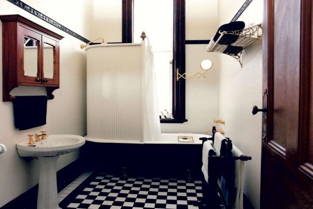 A compact traditional bathroom with a pedestal basin, freestanding bathtub partially screened by a tall radiator, black-and-white checkerboard floor tiles, dark lower wall panelling, white upper walls, ornate trim, a mirrored wall cabinet, and dark timber door and window frames.