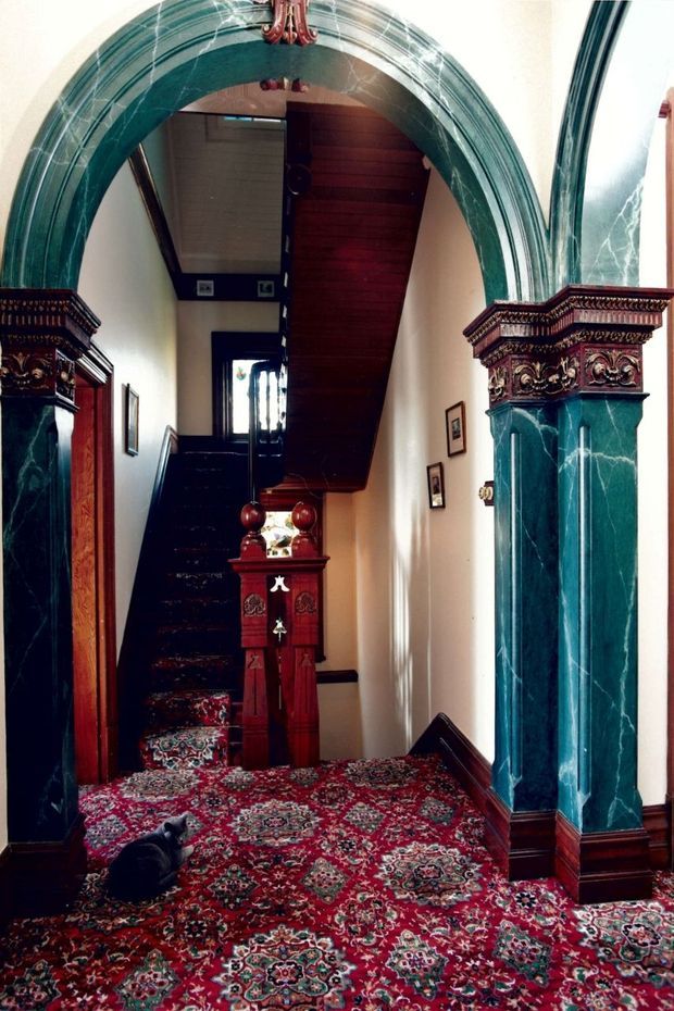 A richly detailed traditional hallway and stairway featuring a grand dark green marble arch and column surround with ornate carved capitals, dark timber wall panelling and staircase, framed artwork, and an elaborate red patterned carpet. A small decorative wooden stand sits at the foot of the stairs, while a cat rests on the floor.