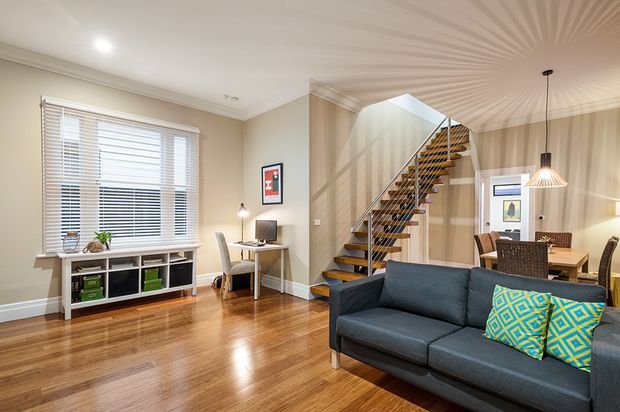A bright open-plan living room with polished timber flooring, beige walls, white plantation blinds, and a dark charcoal sofa with patterned accent cushions. A compact home office nook sits beneath the window, while a timber open-riser staircase with dark metal railings leads upward. A dining area with pendant lighting is visible behind the sofa.