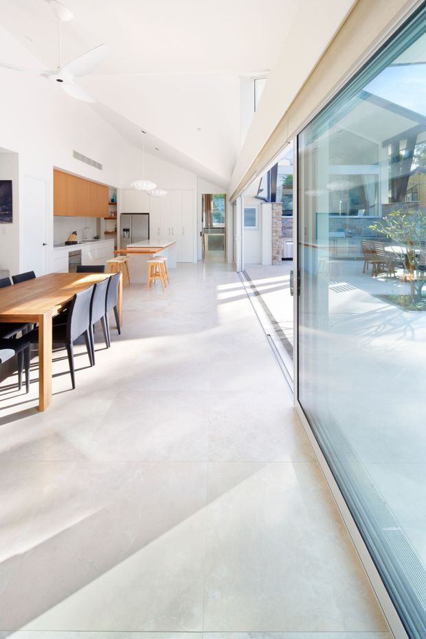 Bright, open-plan contemporary kitchen and dining area with a long timber dining table, black dining chairs, white cabinetry, timber overhead cupboards, and a central island with stools. Full-height glazed sliding doors open the room to a light-filled outdoor courtyard, while a white ceiling fan and pendant lights add functional detail. The space has a clean, minimal material palette and polished pale flooring.