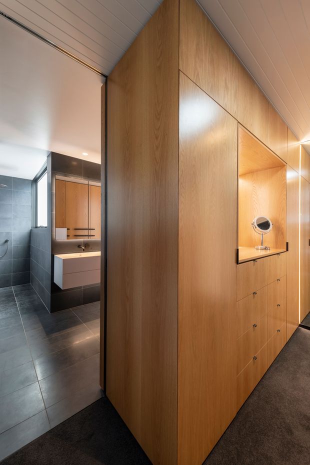 A refined interior view dominated by a full-height timber built-in wardrobe and dressing joinery, with an illuminated recessed vanity and round mirror. To the left, an adjoining dark tiled bathroom is visible with a floating vanity, shower fittings, and grey floor and wall tiles. The space has a warm timber palette, charcoal carpet, clean integrated cabinetry, and a sloped white panelled ceiling.
