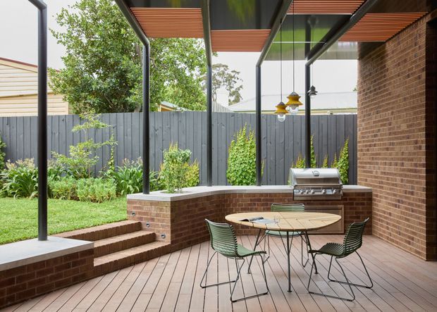 Covered outdoor entertaining area with a timber deck, brick walls and built-in brick barbecue bench. A round outdoor dining table with wire-frame chairs sits beneath a slim contemporary pergola with timber slatted ceiling panels and dark metal posts. Steps lead down to a landscaped lawn and garden enclosed by a dark vertical-board fence.