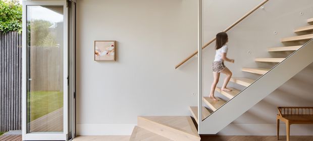 Bright contemporary interior showing an entry area and open staircase. Pale timber stair treads and handrail contrast with white walls, a slim light-coloured balustrade, full-height glazing, and minimalist built-in or adjacent joinery. A child is ascending the stairs, with greenery visible outside through the glass.