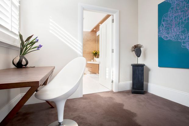 Bright, refined interior view of a compact dining or study area with a sculptural white chair and dark timber table beside a shuttered window. White walls with decorative lower moulding, a taupe carpet, contemporary artwork, and a pedestal display create a polished transitional setting. An open doorway leads to a bright bathroom or dressing area with timber cabinetry and a skylight or timber ceiling detail.