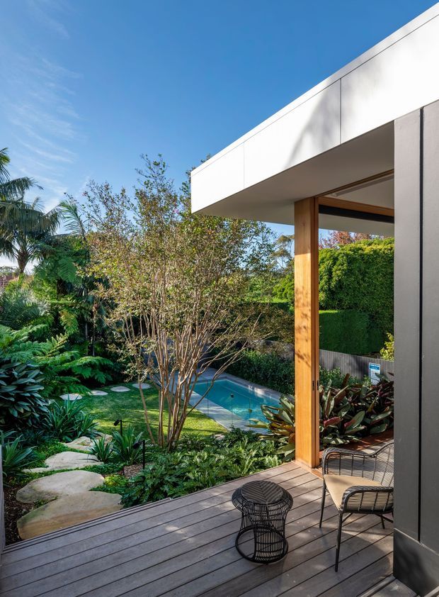 A contemporary outdoor deck beside a modern home, overlooking a lush landscaped garden and compact swimming pool. The scene features a crisp white flat roof and facade, dark-framed glazing, a warm timber post, grey timber decking, and minimalist black wire patio furniture. Tropical and subtropical planting surrounds stepping-stone paths and the pool under a clear blue sky.