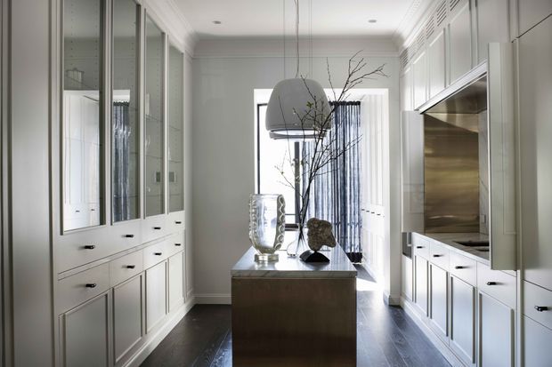 A refined galley-style kitchen with symmetrical white built-in cabinetry, paneled doors and drawers, integrated appliances, dark timber flooring, and a central island with a stone or metallic benchtop. Glass-front cabinetry lines the left wall, while a large sculptural pendant light and a tall branch arrangement create a focal point. Bright daylight enters through the rear glazing, giving the space a polished, contemporary-classic feel.